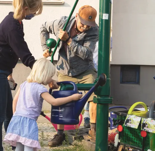 An older man operates a street pump and fills the watering can of a woman and a young girl